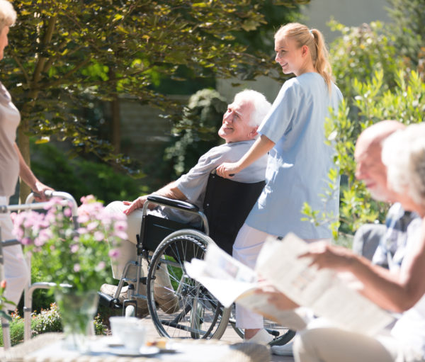 Nurse supporting senior man in wheelchair during meeting with friends in garden on sunny day